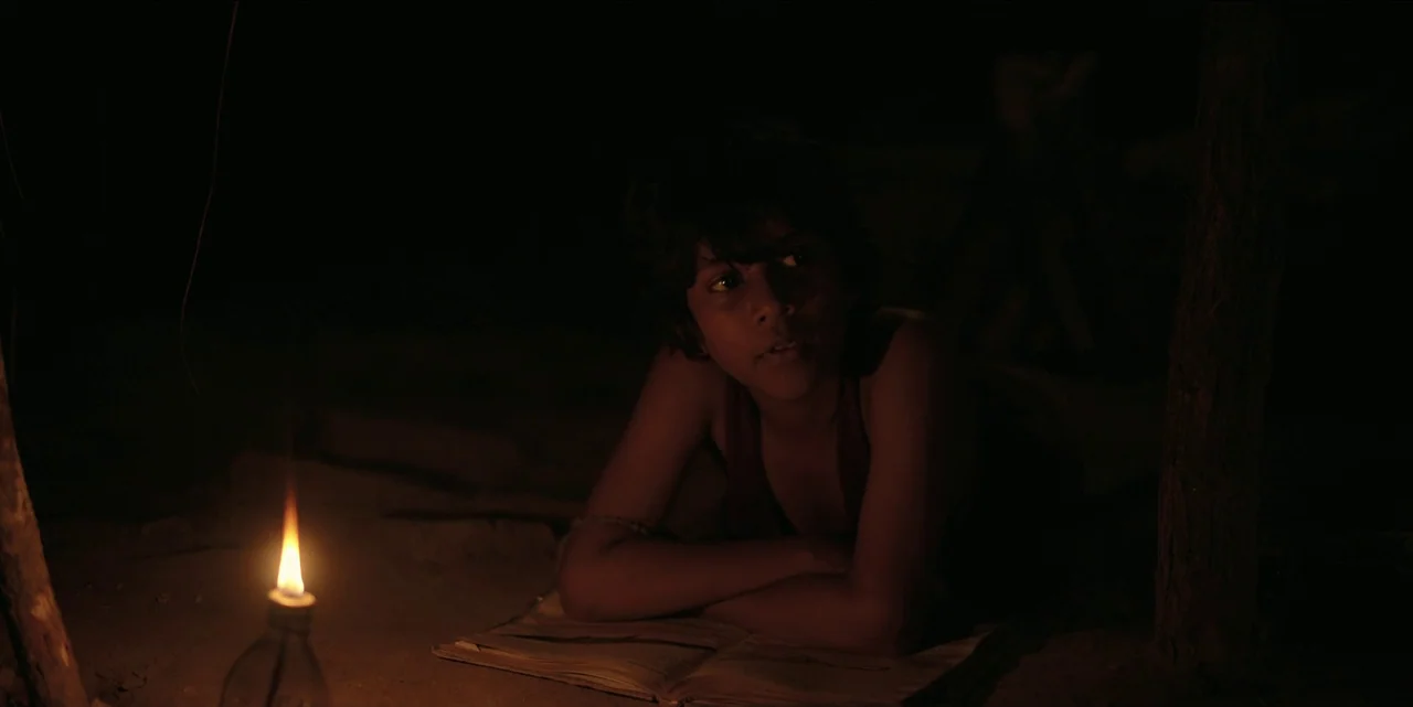 The White Tiger (2021) movie still: medium — A child lies on the dirt ground at night near a small oil lamp. An open book rests on the ground…