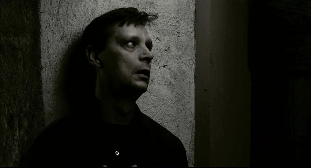 The Secret Village (2013) movie still: closeup — A man stands in a dark corner against a rough concrete wall. The man leans his head against the…