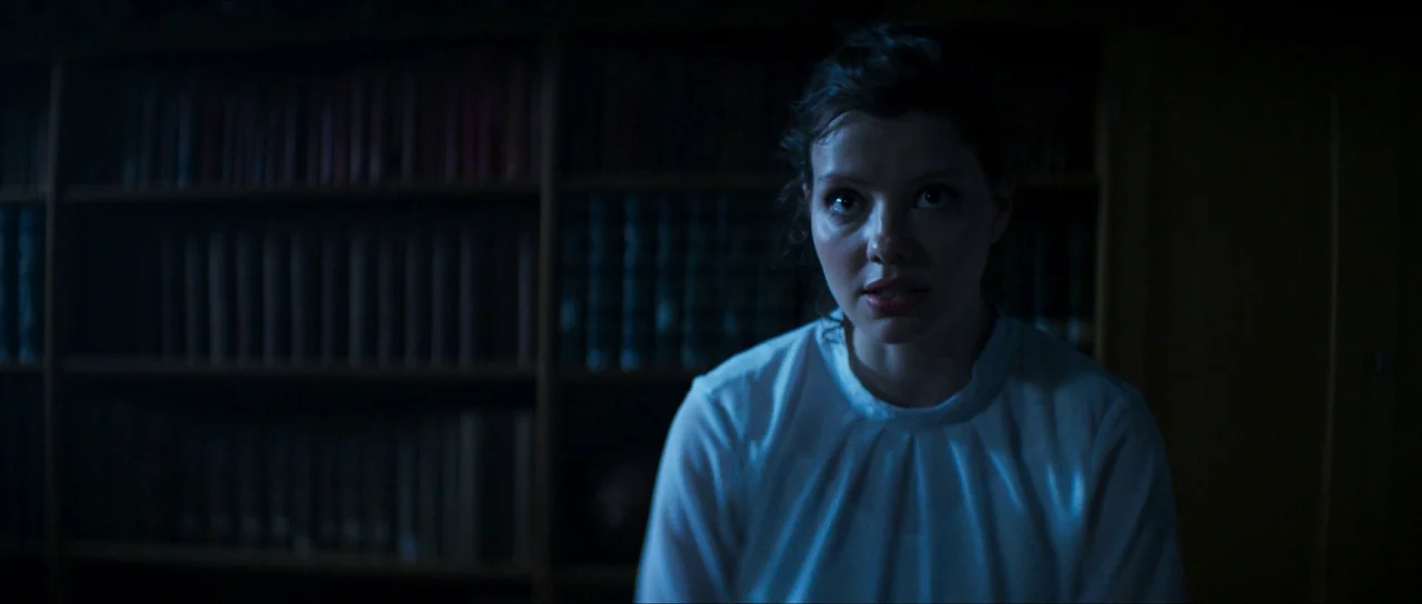 The Sacrifice Game (2023) movie still: medium — A dimly lit room features a wall filled with dark, obscured wooden bookshelves. A young woman in a…