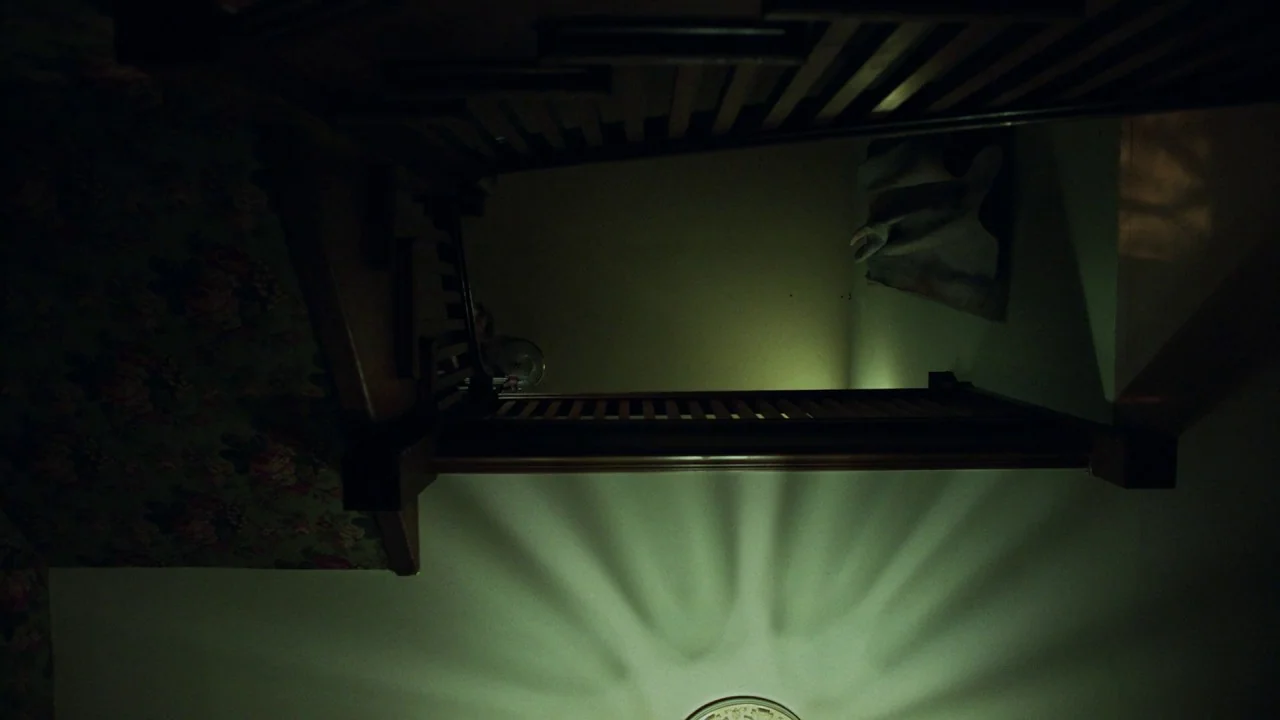 Girl on the Third Floor (2019) movie still: establishing — A dimly lit wooden staircase is viewed from directly above looking down the shaft. Long…