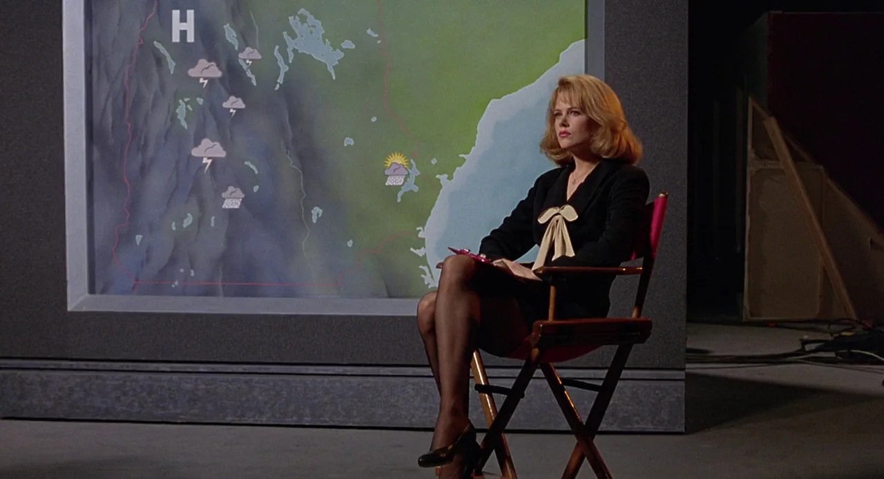 To Die For (1995) movie still: medium — A television news weather studio features a large projected green and gray weather map. A blonde woman in a…
