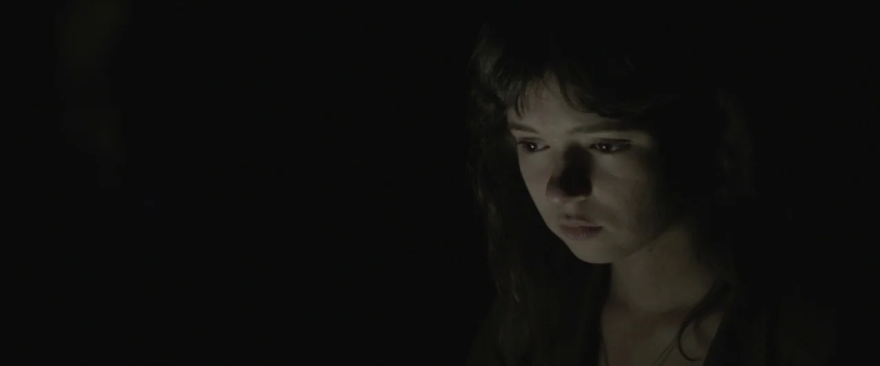 Here Alone movie still: closeup — A person is positioned in a completely dark space with no visible background details. The subject's face is…