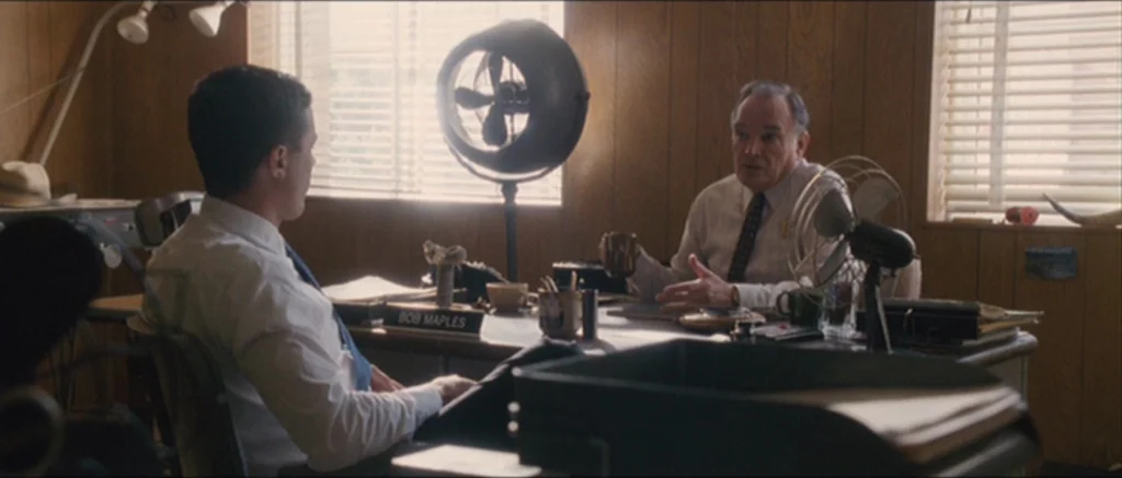 The Killer Inside Me (2010) movie still: medium — A wood-paneled office features a desk with a nameplate, two electric fans, and blinds. Two men are…