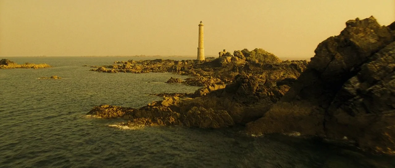 A Very Long Engagement (2004) movie still: establishing — A remote lighthouse sits on a series of jagged rocks surrounded by the sea. The landscape…