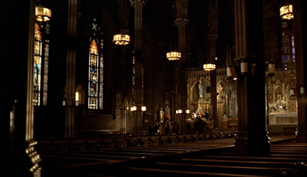The Godfather (1972) movie still: wide — A dark, expansive cathedral nave features rows of wooden pews and stone pillars. Stained glass windows and…