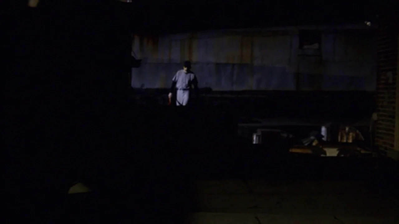 Murder Party movie still: wide — An dark urban exterior space at night with a large brick wall background. A solitary figure stands in a small patch…