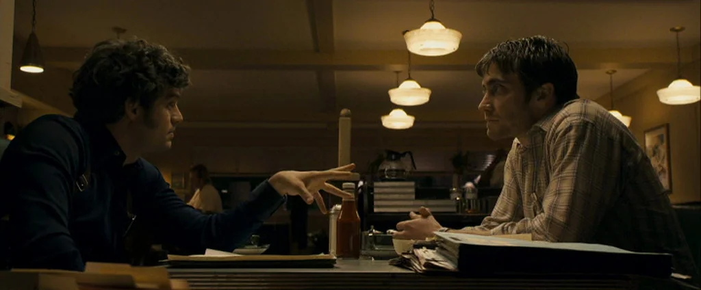 Zodiac (2007) movie still: medium — Two men sit facing each other at a dimly lit booth in a vintage diner. One man leans forward with his hand…