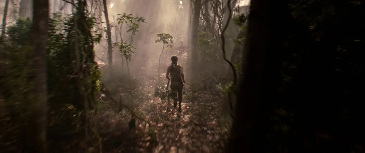 Tomb Raider (2018) movie still: wide — A dense, overgrown jungle with thick fog settling between the tall tree trunks. A woman walks away from the…