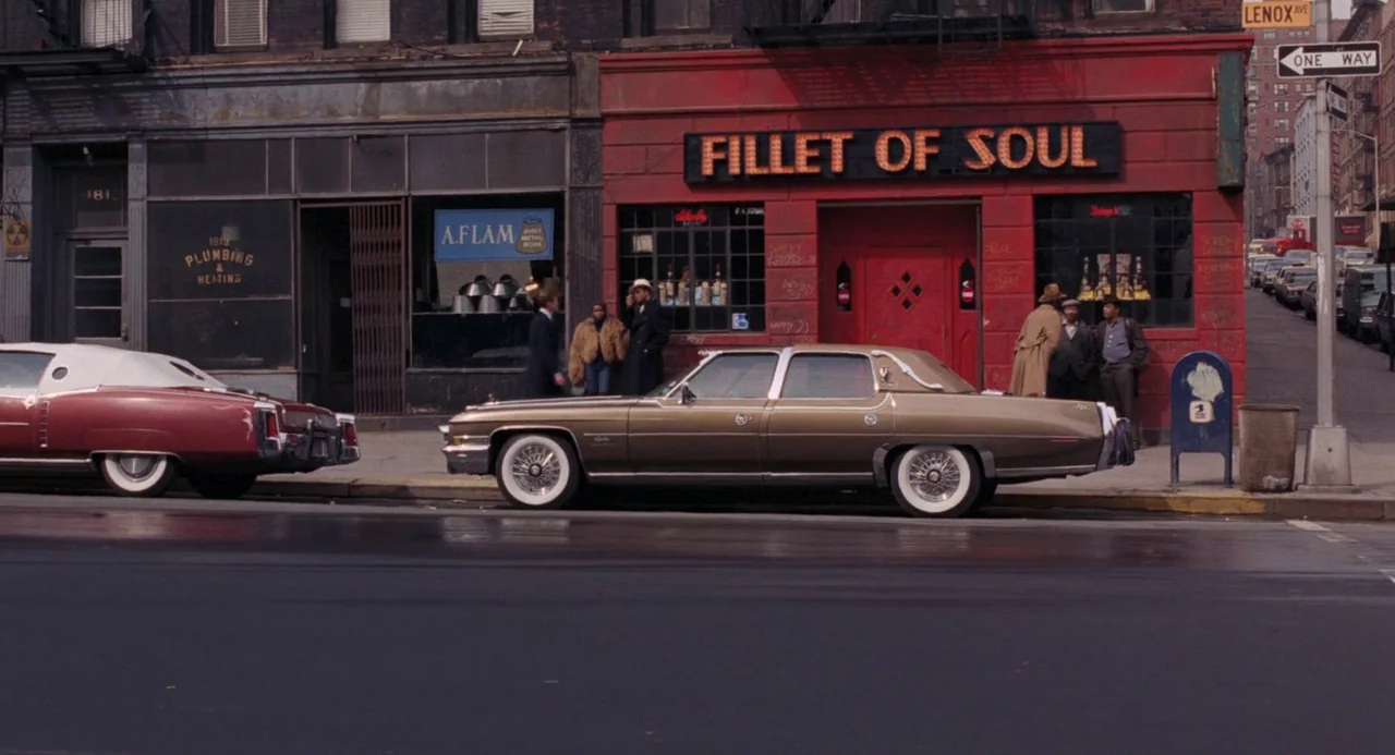 Live and Let Die (1973) movie still: wide — A city street corner features a red building with a Fillet of Soul sign. Several men stand on the…