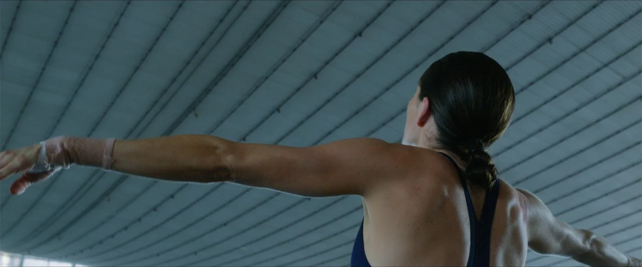 Dive (2013) movie still: medium — An athlete stands inside a large sports arena with a ribbed ceiling overhead. The woman performs an arm stretch…