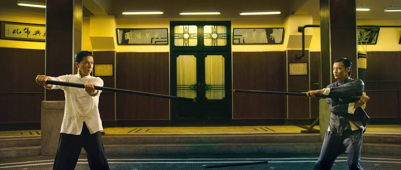 Ip Man 3 (2015) movie still: wide — A spacious interior with wooden wall paneling and rectangular framed Chinese calligraphy signs overhead. Two men…