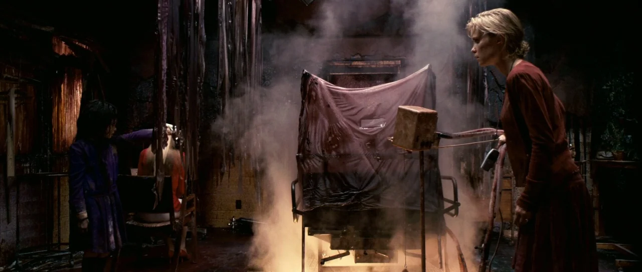 Silent Hill movie still: wide — A dark, dilapidated room with peeling walls and hanging debris contains a smoking metallic chair. A woman stands on…