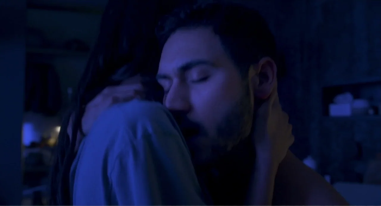 Straight (2024) movie still: closeup — A dimly lit room at night with dark blue ambient lighting. A man and a woman are holding each other in a…