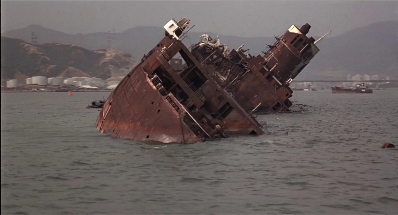 The Man With the Golden Gun (1974) movie still: wide — A heavily rusted, partially sunken ship tilts precariously in a gray, calm harbor. Distant…
