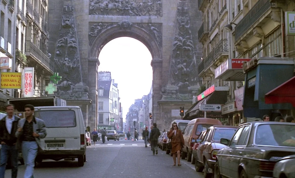 A Tale of Springtime (1990) movie still: wide — A Paris street leads toward the large stone arch of the Porte Saint-Denis. Pedestrians walk along…