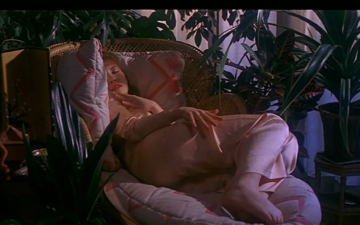 Pepi, Luci, Bom and Other Girls Like Mom (1980) movie still: medium — A woman reclines in a wicker chair surrounded by several large tropical potted…