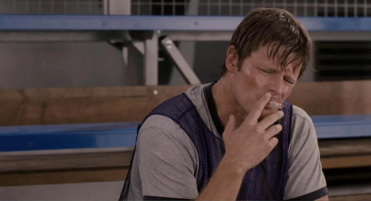 Management (2009) movie still: medium — A set of wooden bleachers with blue metal supports serves as the backdrop. A man wearing a sports mesh…