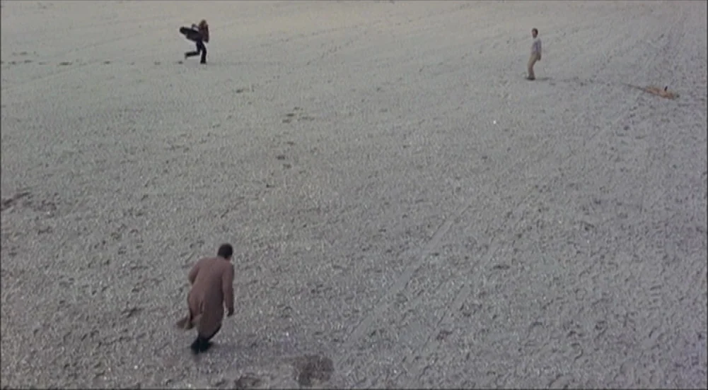 The King Of Marvin Gardens movie still: wide — A vast, empty beach with light gray sand stretches across the entire frame. Three men move across the…