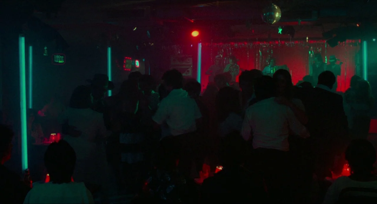 True Stories (1986) movie still: wide — A crowded dimly lit nightclub interior features dancers on the floor in front of a stage. Vertical cyan neon…