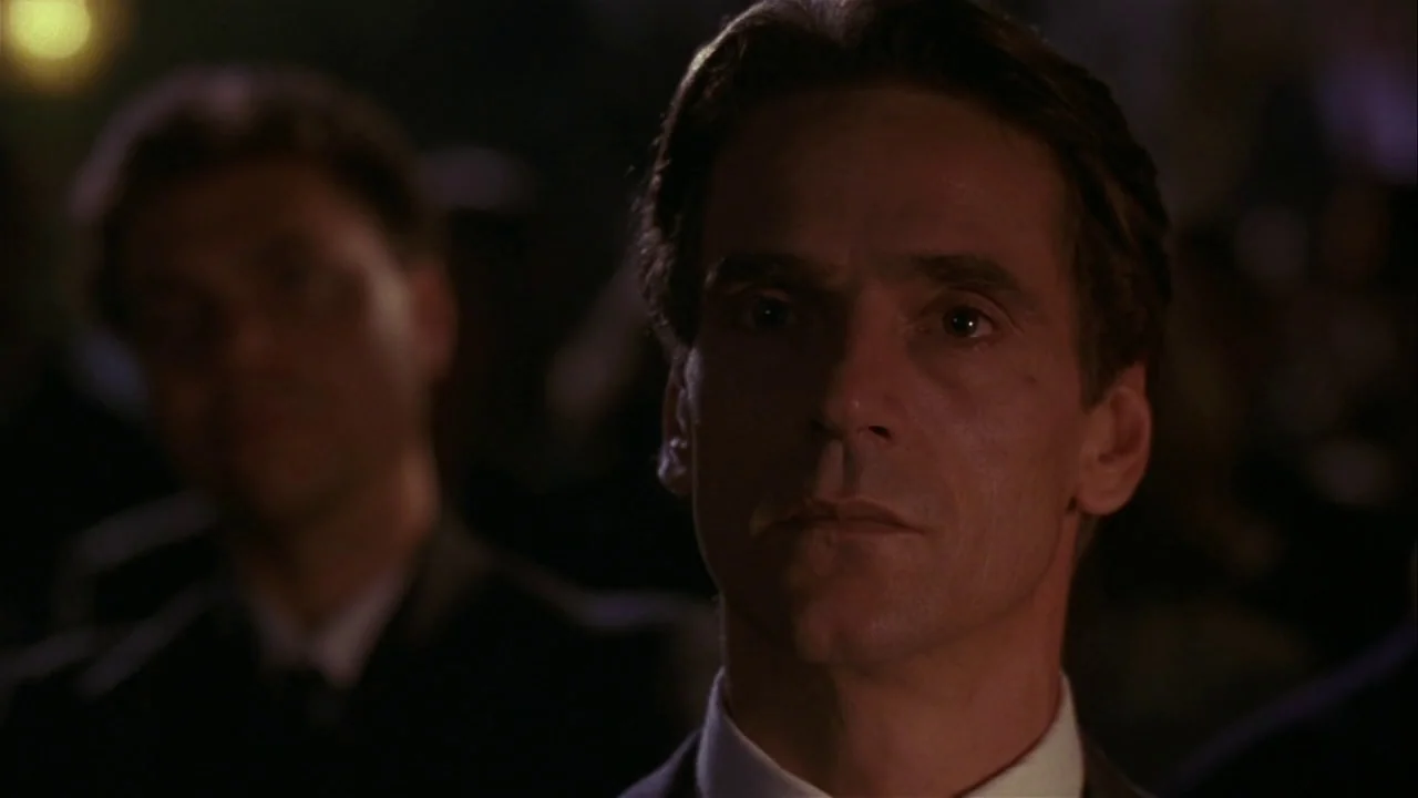 M. Butterfly (1993) movie still: closeup — A dimly lit auditorium setting with rows of seats occupied by indistinct figures. A middle-aged man with…