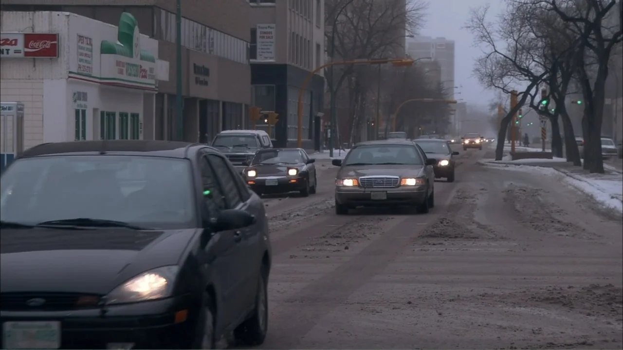 O código oculto (2025) movie still: wide — A snow-covered city street features multiple cars driving through slush on a grey day. Several commercial…