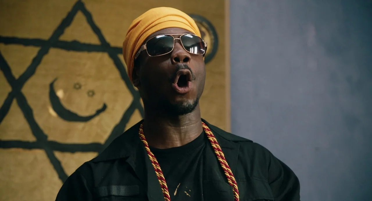 The Day Shall Come (2019) movie still: medium — A room features a large, hand-drawn black Star of David on a gold wall. A man in a yellow durag and…