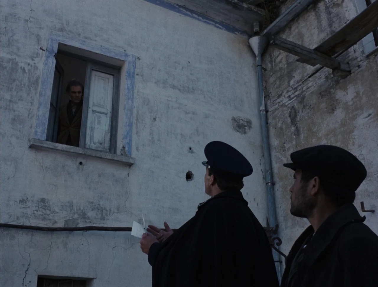 Christ Stopped at Eboli (1979) movie still: wide — A weathered, light-colored stucco exterior wall with a high window and a metal drainpipe. Two men…