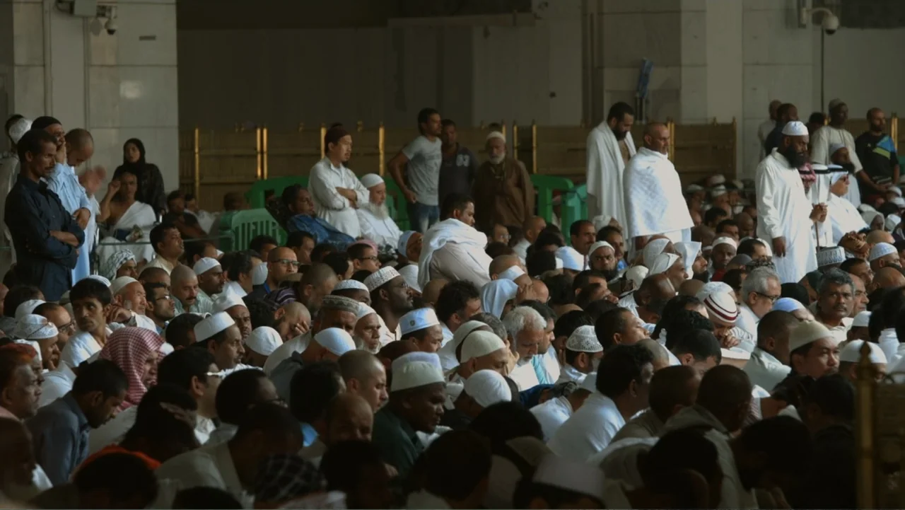 One Day in the Haram (2017) movie still: wide — A spacious hall with marble columns and white walls houses a dense gathering. Hundreds of men sit on…