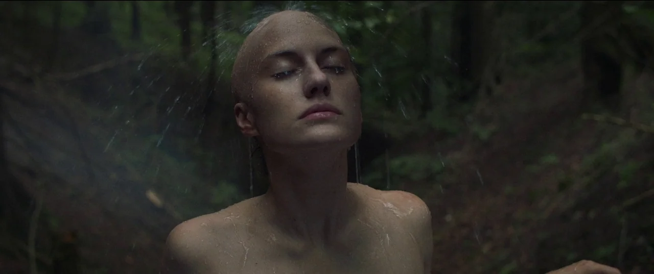 Nightsiren (2022) movie still: medium — A dense forest with blurred trees fills the dark, verdant background. A woman with a shaved head stands in…