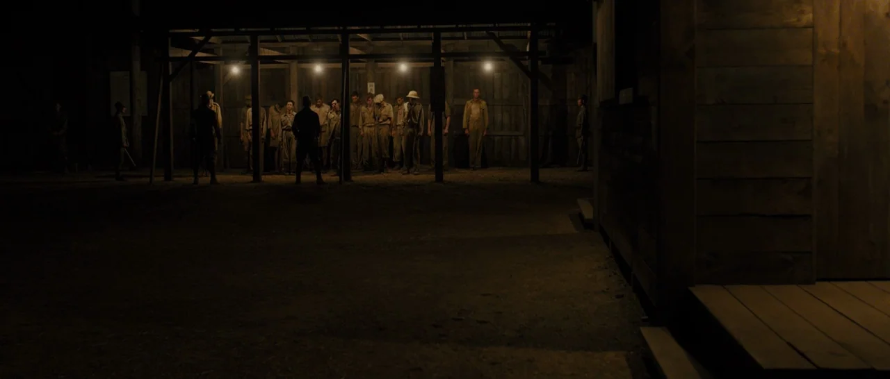 Unbroken (2014) movie still: wide — A dark, dirt-floored military encampment features men standing in rows under a covered structure. Several men in…