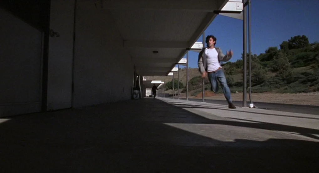 Brick (2005) movie still: fullbody — An outdoor concrete corridor runs alongside a long building under a clear blue sky. A man runs towards the…