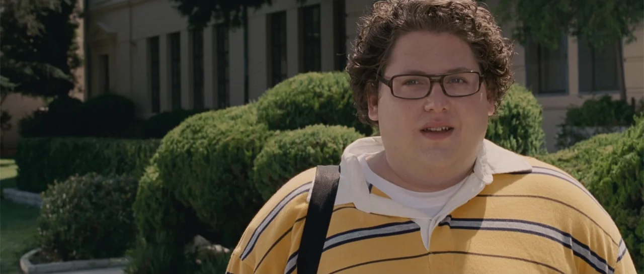Accepted (2006) movie still: medium — A college campus exterior with manicured hedges and a building in the background. A young man in a striped…