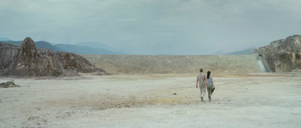 Blackhat (2015) movie still: wide — A wide expanse of dry, barren earth stretches toward a massive industrial dam. A man and a woman walk away from…
