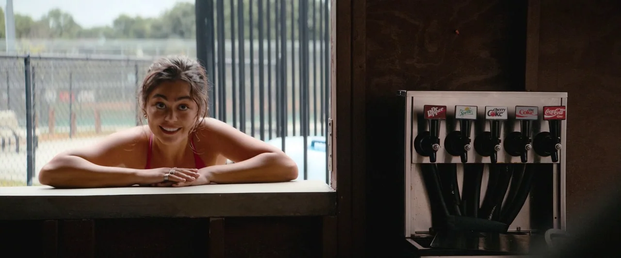 Snack Shack (2024) movie still: medium — A wooden concession stand counter faces an outdoor area enclosed by a metal fence. A young woman smiles…