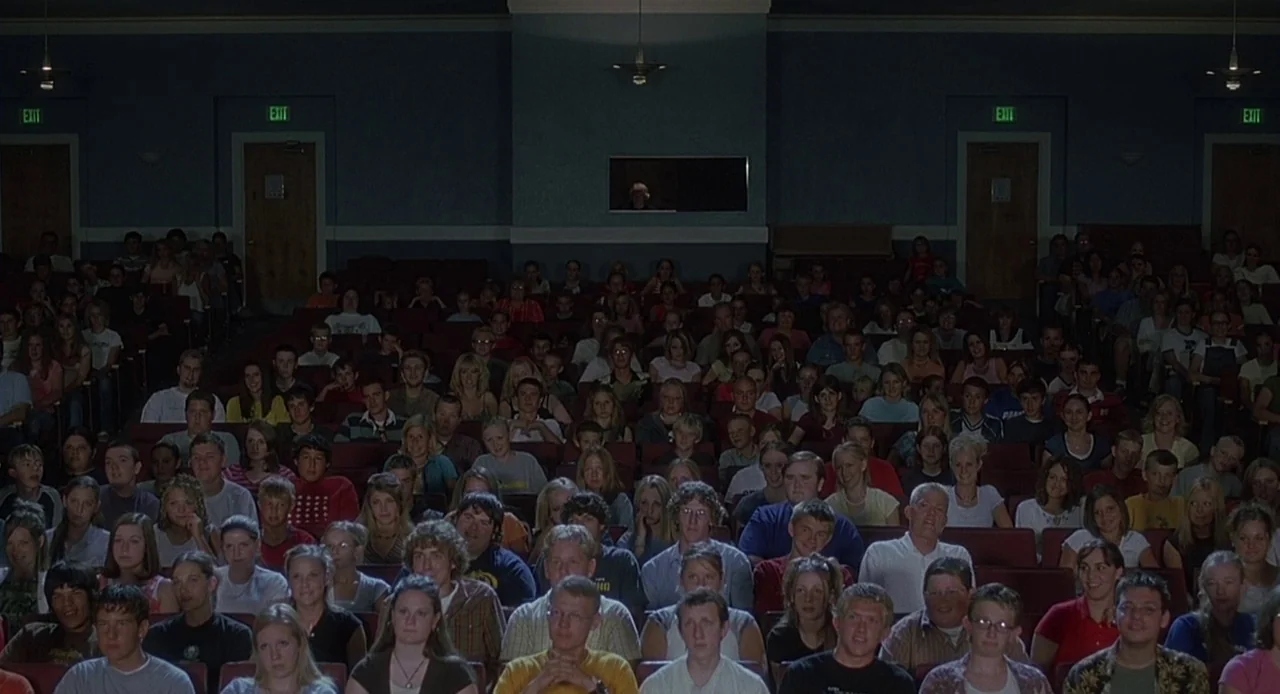 Napoleon Dynamite (2004) movie still: wide — A crowded auditorium features rows of red velvet seats filled with people facing forward. The room is…