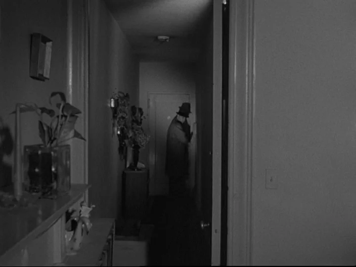 Blast Of Silence (1961) movie still: wide — A dark, narrow residential corridor features walls with a plant and decorative items. A man in a hat…