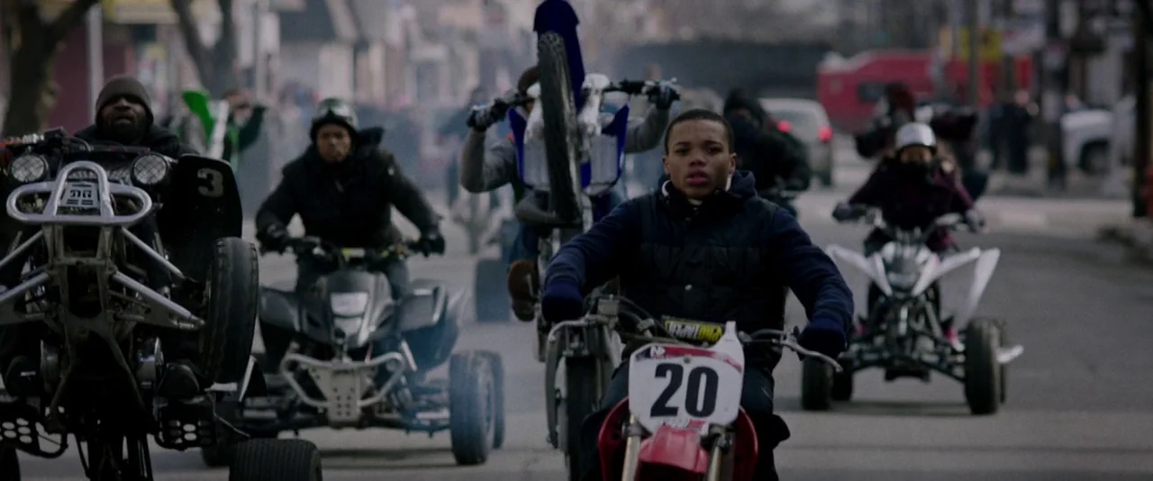 Creed (2015) movie still: wide — An urban neighborhood street is filled with people riding quad bikes and dirt bikes. A young man in a dark vest…