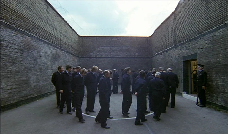 A Clockwork Orange (1971) movie still: wide — A high-walled concrete exercise yard has barbed wire running along the top edges. A group of men in…
