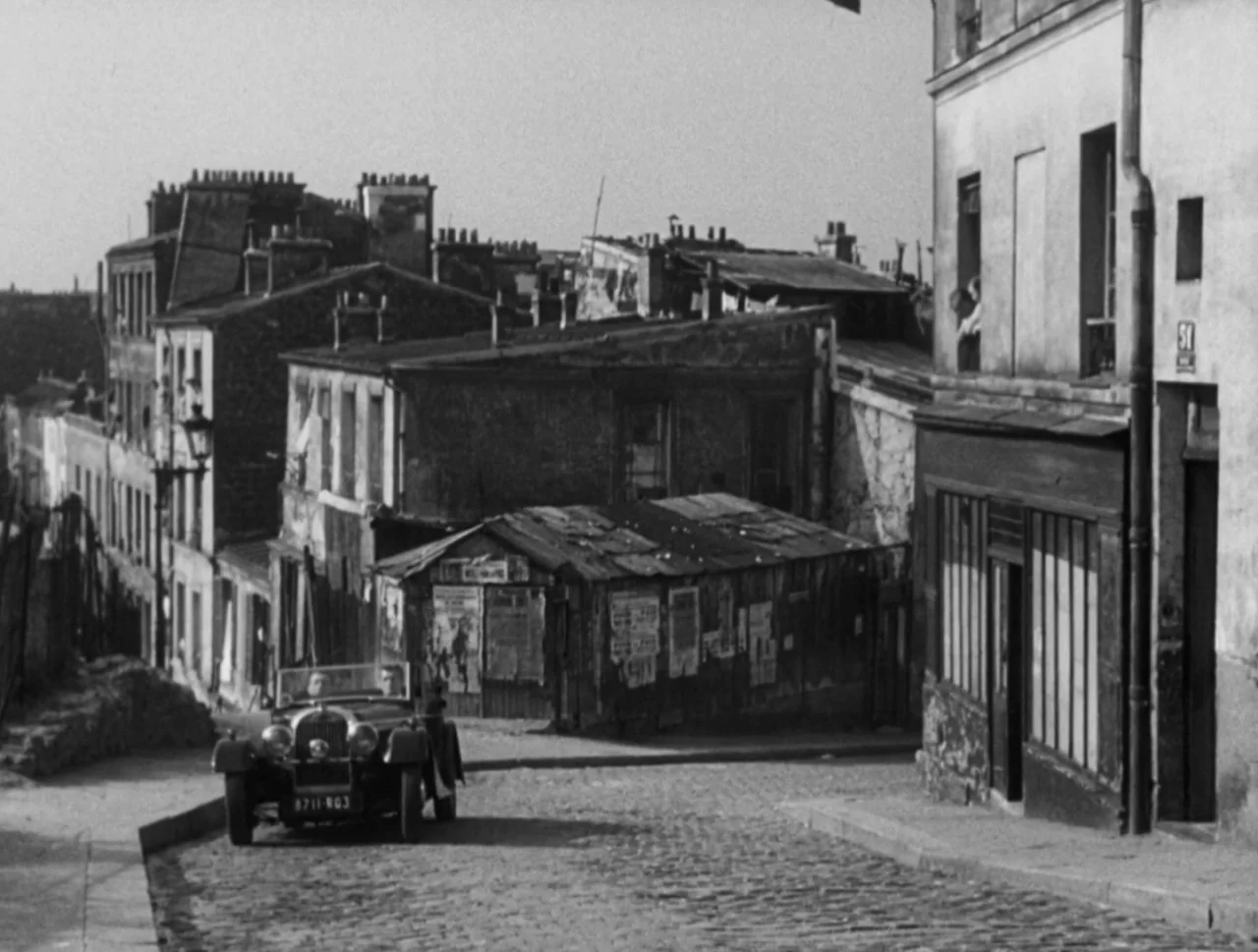 Orpheus (1950) movie still: wide — A narrow European cobblestone street winds through a dense area of old urban buildings. A vintage convertible car…