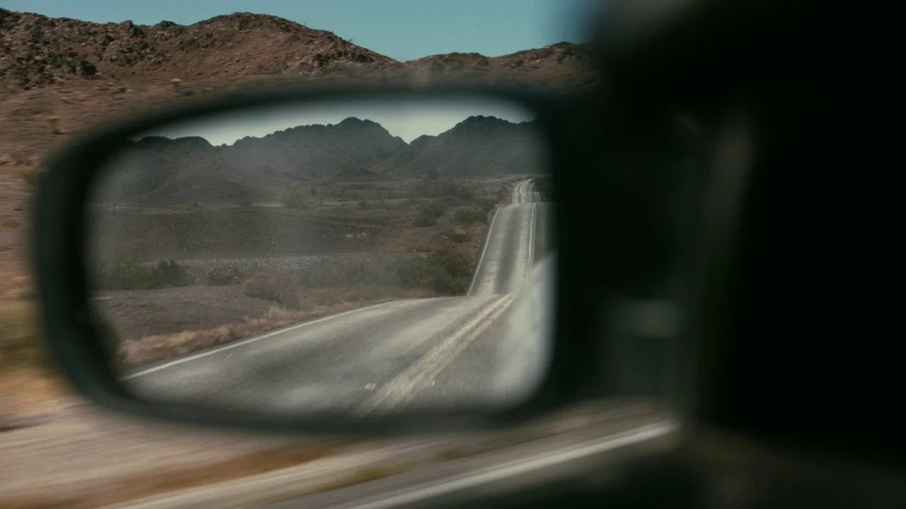 One Battle After Another (2025) movie still: closeup — A side-view car mirror captures a desert road winding toward distant rocky mountains. The…