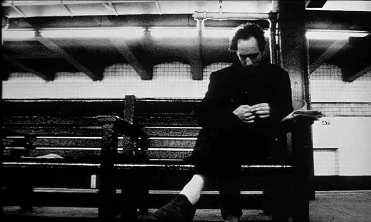 Pi (1998) movie still: medium — A dark, empty subway platform features tiled walls and heavy wooden seating. A lone man sits on the bench while…