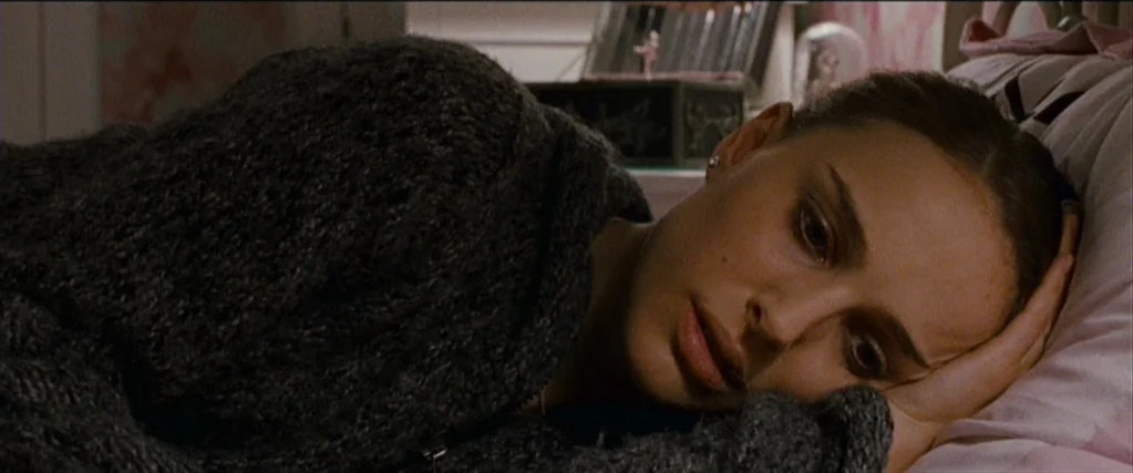Black Swan (2010) movie still: closeup — A dimly lit space features pink wall patterns and a decorative music box. A young woman rests on a bed,…