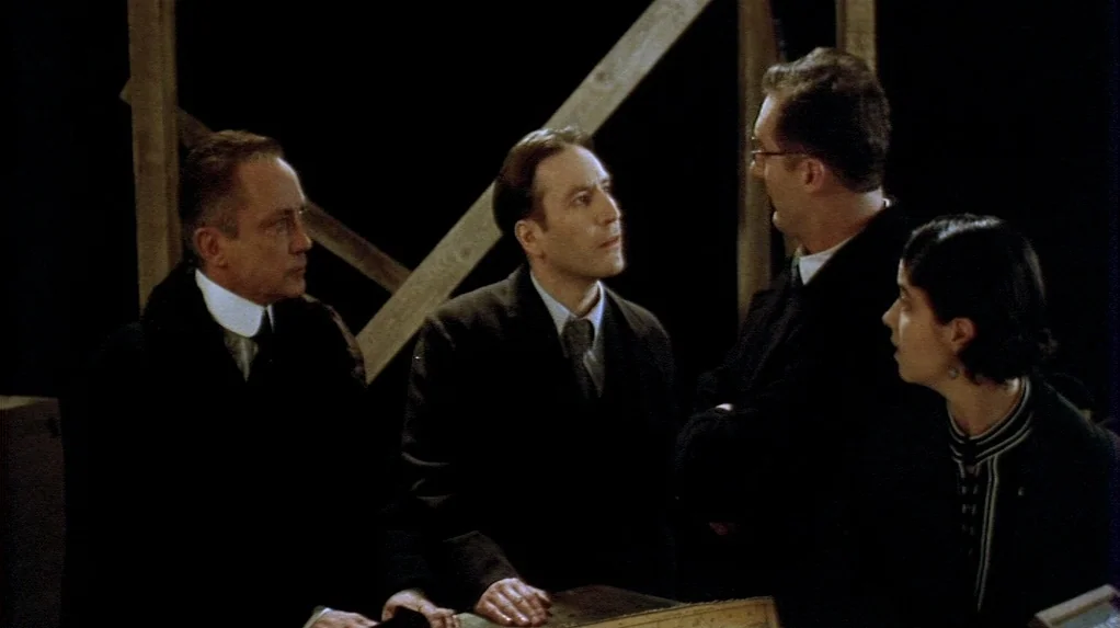 Shadow of the Vampire (2000) movie still: medium — A group of four people stands around a surface within a dark room. The backdrop is sparse with…