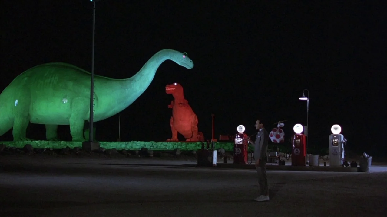 Pee-Wee’s Big Adventure (1985) movie still: wide — A nighttime roadside area features a large green brontosaurus statue and a smaller red t-rex. A…