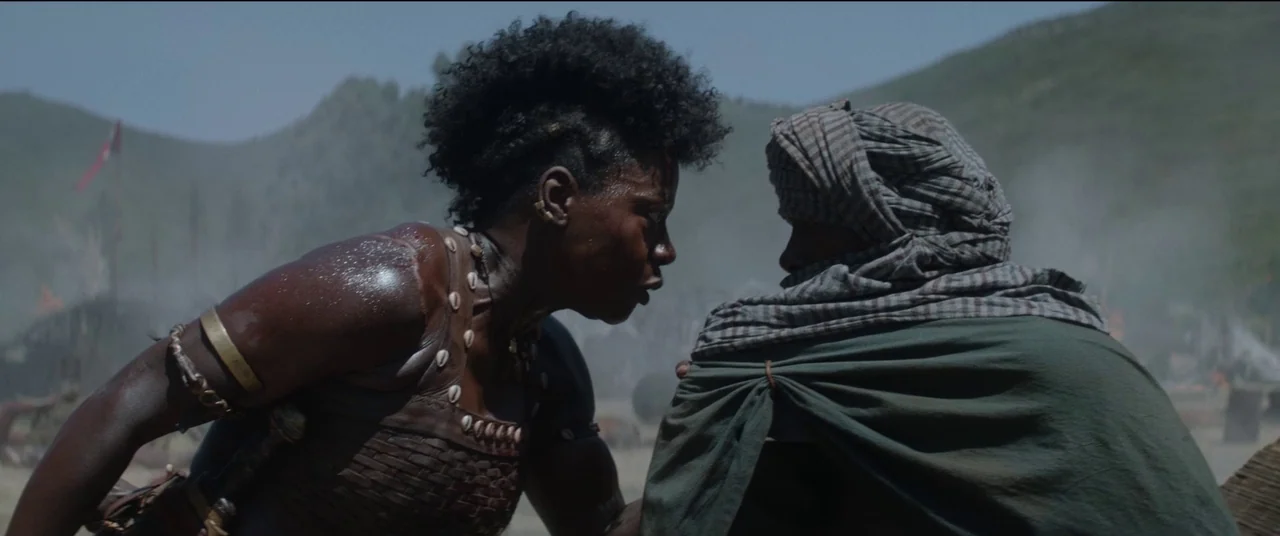 The Woman King (2022) movie still: medium — A dusty outdoor battlefield features rolling hills and smoldering debris in the background. A woman in…