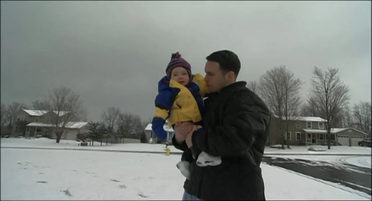 Hard as Nails (2001) movie still: medium — A snow-covered suburban street features houses and bare trees under a cloudy sky. A man stands in the…