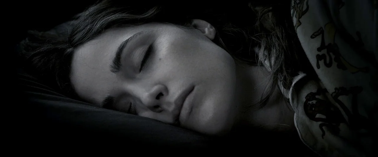 Insidious (2010) movie still: closeup — A close view shows a woman sleeping in a dark environment. Her face rests against a pillow with a patterned…