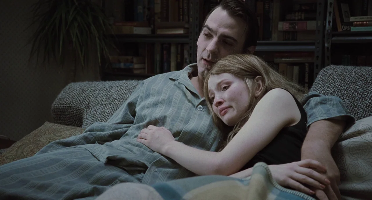 Sleeping Beauty (2011) movie still: medium — A dimly lit room features a bookshelf filled with books in the background. A man in pajamas sits on a…