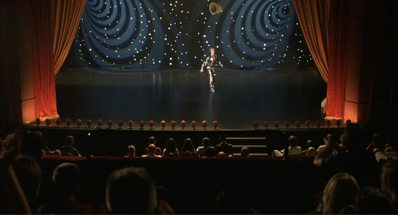 Scoop (2024) movie still: wide — A dark, occupied auditorium looks toward a brightly lit performance stage with red curtains. A solitary performer…