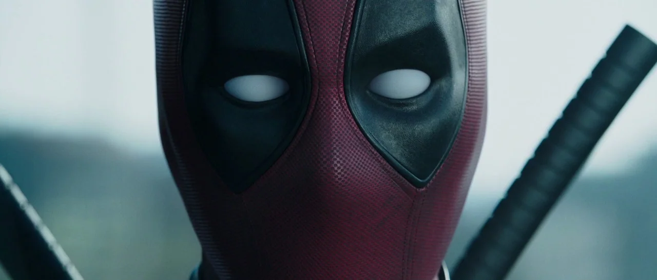 Deadpool (2016) movie still: closeup — A character wears a textured red and black mask with opaque white eye lenses. Two sword handles protrude…